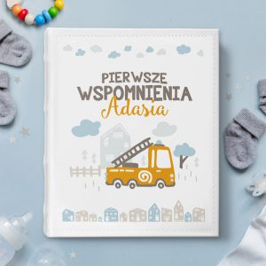 Album pamiątkowy na pierwsze zdjęcia dziecka. Na białej okładce jest rysunek żółtego wozu strażackiego na tle domowego zacisza. Na górze personalizowany nadruk pierwsze wspomnienia z imieniem Waszego synka.