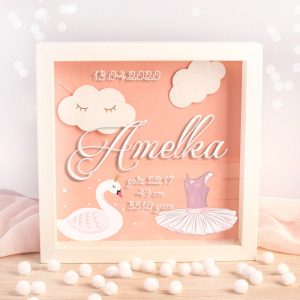 Kwadratowa metryczka 3D z białą, drewnianą ramką. Z przodu jest przezroczysta szybka z trwałym nadrukiem - imieniem dziecka i metryką urodzenia. Na różowym tle wiszące chmurki, łabędź i sukienka baletnicy.