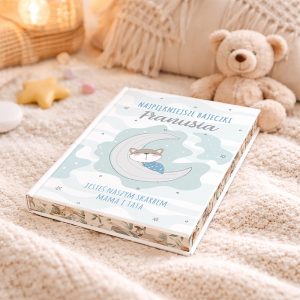 Ilustracja książki "Najpiękniejsze Bajeczki Franusiu" z rysunkiem kota śpiącego na księżycu, umieszczona na miękkim pledzie obok pluszowego misia oraz dekoracji w pastelowych kolorach.