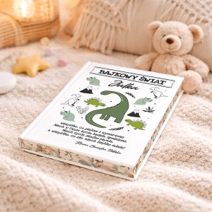 Książka zatytułowana "Bajkowy Świat Julka", leżąca na miękkim, jasnym pledzie, obok pluszowego misia. Okładka książki zdobiona ilustracjami dinozaurów i zwierząt, z zielonym dinozaurem jako centralnym motywem.