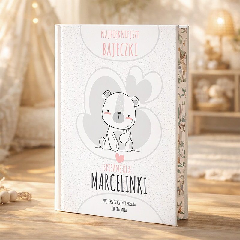 Otwarta książka z tytułem 'Najpiękniejsze bajeczki' na okładce, przedstawiająca uśmiechniętego misia w szaro-białych odcieniach, z sercem w dolnej części i napisem 'Spisane dla Marcelinki' oraz 'Najlepsze życzenia składam, ciocia Ania'. W tle widać przytulne wnętrze z rozmytymi meblami i jasnym oświetleniem.