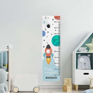 Miarka wzrostu to dekoracja wisząca do pokoju dziecka. Wzbogacona jest o grafikę, która przedstawia motyw kosmiczny z rakietą kosmiczną, astronautą i planetami.
