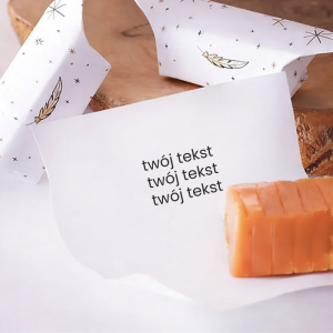 Kawałek pomarańczowego sera na białym papierze z napisem „twój tekst”, obok opakowania w subtelnym wzorze.