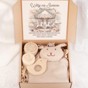 Zestaw powitalny dla dziecka w kartonowym opakowaniu, zawierający miękkiego zajączka, drewniane gryzaki w kształcie ptaszka i dwa okrągłe pojemniki z motywami zwierzęcymi oraz kartkę z życzeniami na tle karuzeli z konikami.