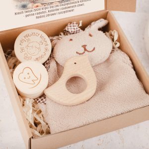 Pudełko prezentowe zawierające miękką zabawkę w kształcie królika, drewniane gryzaki w kształcie ptaka i zarysowanej buzi oraz dwa okrągłe pojemniki z wygrawerowanymi napisami, na tle ścinki wypełnionej sianem.