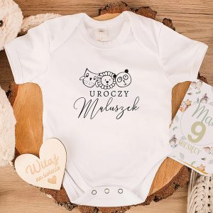Biały body dla niemowlaka z napisem 'Uroczy Maluszek' oraz rysunkami zwierząt: psa, lwa i pandy, na tle naturalnego drewna i pluszowego misia. Obok znajduje się kartka z napisem 'Witaj na świecie' i książeczka o miesiącach.