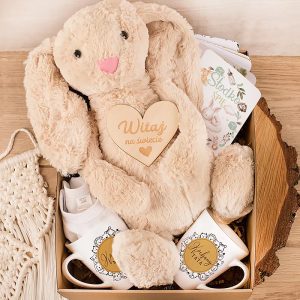 Zestaw prezentowy zawierający pluszowego królika z serduszkiem 'Witaj na świecie', dwie białe filiżanki z napisami, małe skarpetki, dekoracyjną kartkę oraz fragment makramy w tło z naturalnego drewna.