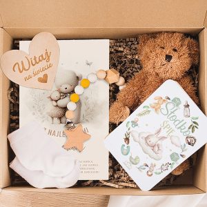 Zestaw urodzinowy dla dziecka w kartonowym pudełku, zawierający przytulankę w kształcie misia, drewniany smoczek, białe skarpetki, kartkę powitalną z kolorową grafiką zwierząt oraz książeczkę dla dzieci z ilustracjami zająca i innych postaci, wszystko na wypełnieniu z papierowej wiórówki.