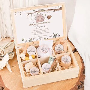 Drewniana skrzynka z napisem powitalnym dla nowonarodzonego dziecka, zawierająca różne drobiazgi, takie jak ozdobne cukierki, kołysanki i banknot dwuzłotowy, ułożone na słomkowym wypełnieniu.