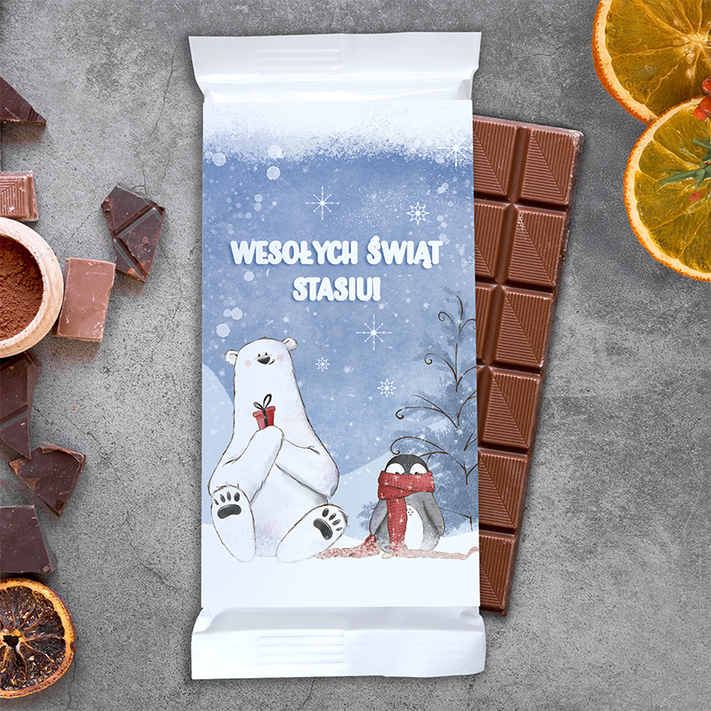 Opakowanie czekolady z ilustracją białego niedźwiedzia trzymającego prezent i pingwina w czerwonym szaliku, na tle zimowego krajobrazu. Na górze napis "Wesołych Świąt Stasiu!". W otoczeniu czekolady znajdują się kawałki ciemnej czekolady, kawałek suszonego owocu i miseczka z kakao.