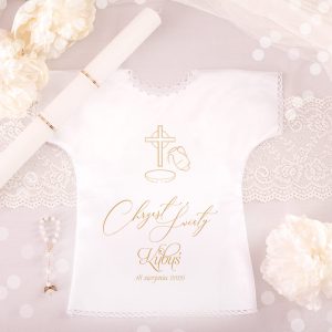 Biała szatka do chrztu z nadrukiem, zawierająca krzyż, dzbanek i obrączkę; podpis "Chrzest Święty Kubuś 18 sierpnia 2025"; otoczenie z białymi kwiatami i krawędzią koronkową.