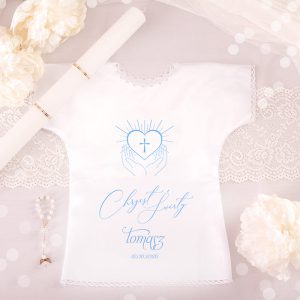 Biała szata z nadrukiem przedstawiającym serce z krzyżem i dłońmi, z napisem "Chrzest Święty Tomasz 12.06.2026". Obok znajduje się biała świeca, różańce, koronkowa koronka i sztuczne kwiaty na tle szaro-biały w kropki.