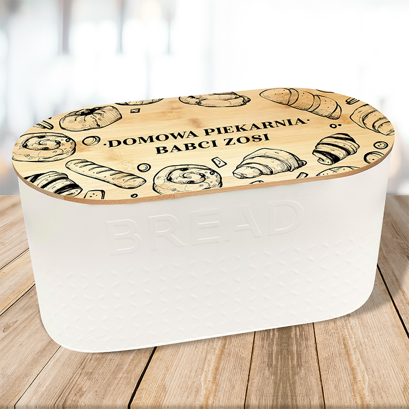 Pojemnik na chleb z białym korpusem i drewnianą pokrywą ozdobioną rysunkami różnych wypieków, z napisem 'DOMOWA PIEKARNIA BABCI ZOSI'. Podstawka z naturalnego drewna na tle ciemnego drewnianego stołu.
