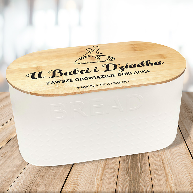 Słoik na chleb z drewnianą pokrywką, na której znajduje się napis 'U Babci i Dziadka Zawsze Obowiązuje Dokładka' oraz imiona 'Wnuczka Ania | Radek'. Słoik ma biały, lekko wypukły korpus z teksturą i znajduje się na drewnianym blacie.
