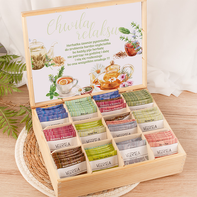 Drewniana skrzynka z różnymi rodzajami herbat w kolorowych woreczkach, z napisem 'Chwila relaksu' na górnej części wieka, otoczona roślinnością na jasnym tle.