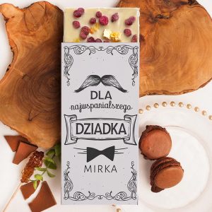 Czekolada z napisem "DLA najwspanialszego DZIADKA MIRKA" na szarym tle, ozdobiona orzechami i owocami, umieszczona na drewnianym tle obok deserów, w tym makaroników i czekolady.