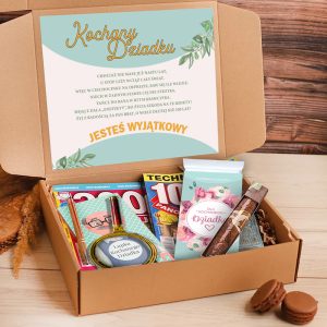 Otwarte pudełko prezentowe z napisem 'Kochany Dziadku' na górnej kartce, wypełnione różnymi drobiazgami, takimi jak słodycze, gazetki i lusterko z rysunkiem dziadka; w tle widoczne makaroniki na stole.