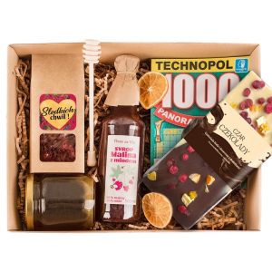 Zestaw prezentowy zawierający syrop malinowy z miodem, miód w słoiku, kawałki czekolady z dodatkami, herbatę ziołową oraz instrukcję do gry planszowej, wszystko ułożone w tekturowym pudełku wypełnionym papierowymi wiórami.