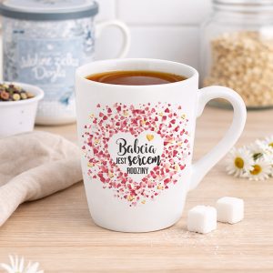 Kubek z napisem "Babcia jest sercem rodziny", ozdobiony kolorowym sercem z płatków, stoi na drewnianym blacie obok cukru i kwiatów. W tle znajduje się słoik z płatkami i ozdobny pojemnik.