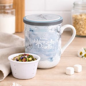 Ceramiczny kubek z pokrywką w błękitnych odcieniach, z napisem "ulubione zioła Dziadka", obok niego mały biały kubeczek wypełniony mieszanką ziół. W tle widoczne są składniki do przygotowania herbaty oraz kostki cukru.