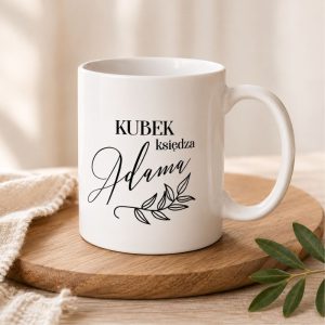 Kubek z napisem "Kubek księdza Adama" na białym tle, ozdobiony liśćmi, stojący na drewnianym talerzu.