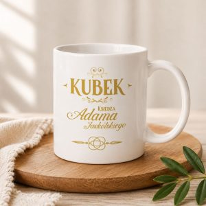 Biały kubek z złotym napisem "KUBEK Księdza Adama Jaskólskiego" na jasnym tle, znajdujący się na drewnianym talerzu.