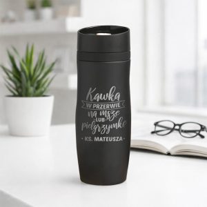 Czarny kubek termiczny z napisem „Kawka w przerwie na nasze pielgrzymkę. KS. Mateusza” na tle biura z roślinami.