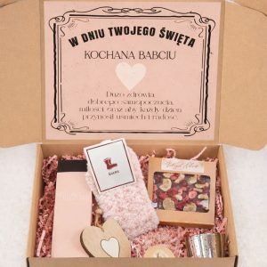 Otwarta paczka z prezentem dla babci, zawierająca różowe skarpetki, kosmetyk Dream, świeczkę, ozdobne serce, oraz opakowanie z suszonymi kwiatami, z napisem życzeń na górze.