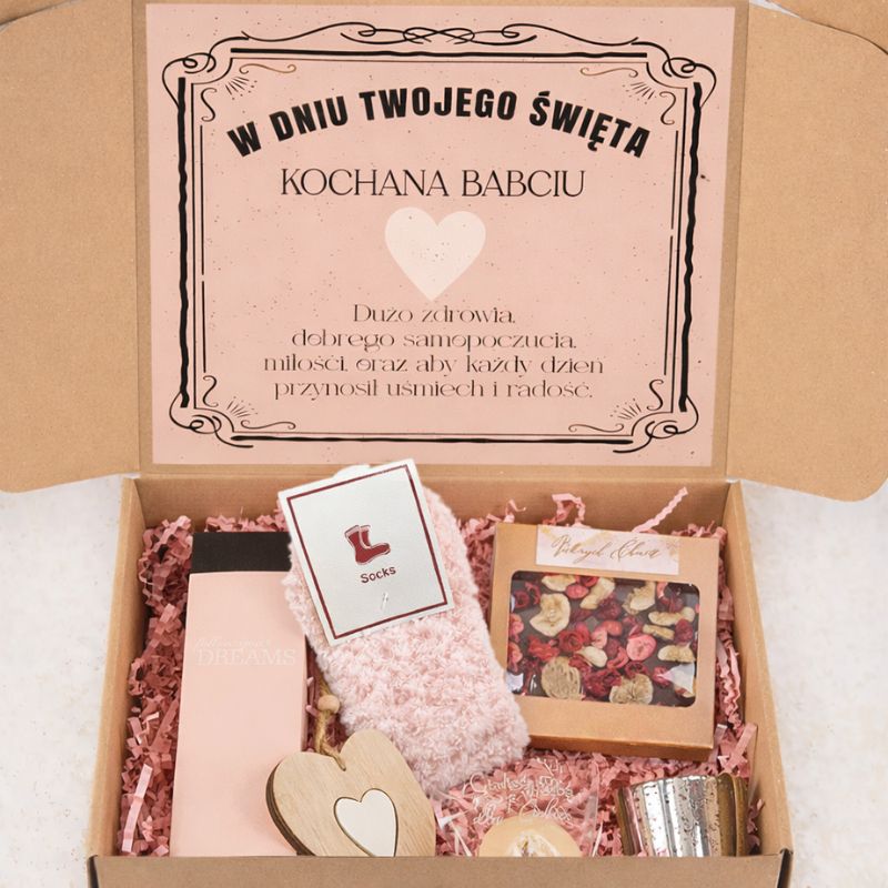 Otwarta paczka z prezentem dla babci, zawierająca różowe skarpetki, kosmetyk Dream, świeczkę, ozdobne serce, oraz opakowanie z suszonymi kwiatami, z napisem życzeń na górze.