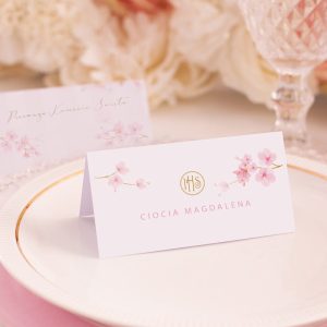 Prostokątna wizytówka z kwiatami różowej magnolii po dwóch stronach. Z jednej widnieje napis "Pierwsza Komunia Święta", a z drugiej imię i nazwisko gościa. Czcionka wykonana jest w eleganckim i ozdobnym stylu.