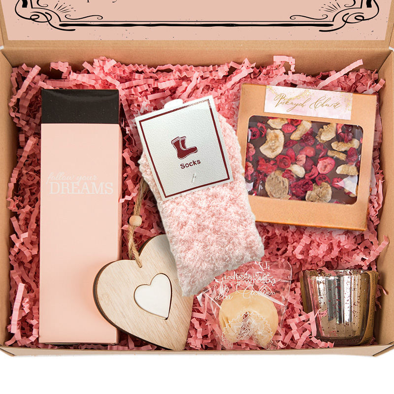 Zestaw prezentowy w pudełku, wypełniony różowym papierem do pakowania, zawierający różowe skarpety, świecę w metalowym pojemniku, drewniane serce z białym wypełnieniem, słodkości i opakowanie z mieszanką suszonych kwiatów. Na jednym z elementów widnieje napis 'follow your DREAMS'.