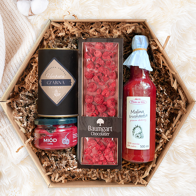 Pudełko w kształcie heksagonu wypełnione produktami spożywczymi, w tym czarna herbata, przetwór owocowy z malin i truskawek, miód oraz pralinki czekoladowe z malinami, wszystkie estetycznie ułożone na papierze wypełniającym.