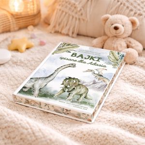 Książka z ilustracjami dinozaurów, zatytułowana "Bajki spisane dla Adasia", leży na miękkim kocu obok pluszowego misia, w otoczeniu dekoracyjnych przedmiotów takich jak gwiazdka i kolorowe cukierki.