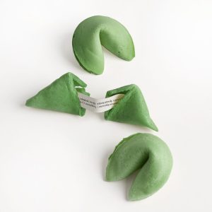 Zielone ciastka z wróżbą, w tym jedno otwarte z widoczną karteczką wewnątrz, na białym tle.