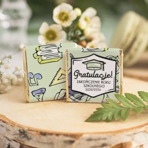 Dwa małe opakowania czekoladek z kolorowym wzorem przyborów szkolnych oraz napisem 'Gratulacje! Zakończenie roku szkolnego 2025/2026', umieszczone na deserowej powierzchni, w otoczeniu kwiatów i roślinności.