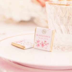 Kwadratowa czekoladka belgijska, zamknięta w złoty papierek. Na wierzchu jest biała etykieta z motywem kwiatów różowej magnolii oraz personalizacja - imię dziecka i data uroczystości.