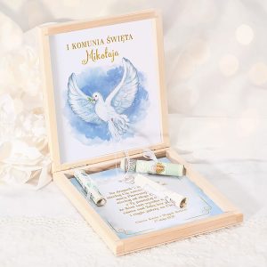 Otwarta drewniana skrzynka z ilustracją białej gołębicy na niebieskim tle oraz kilkoma zwiniętymi papierami w środku, zawierającymi tekst dotyczący I Komunii Świętej, z goldową czcionką.