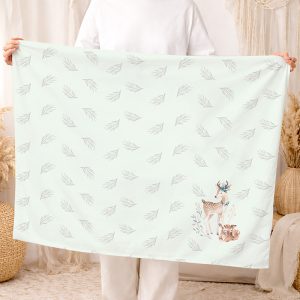 Osoba trzyma dużą, jasnozieloną chustę z delikatnym wzorem liści oraz ilustracjami jelenia i lisa w dolnym rogu.