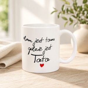 Biały kubek z czarnym napisem "dom jest tam, gdzie jest Tata" oraz małym serduszkiem, ustawiony na stole obok rośliny.