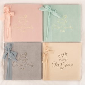 Cztery eleganckie albumy w pastelowych kolorach: różowym, miętowym, szarym i jasnobeżowym, każdy ozdobiony wstążką i napisem 'Chrzest Święty Neli' z obrazkiem huśtawki na okładce.