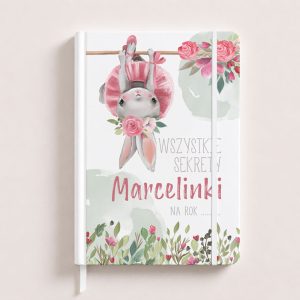 Notatnik z ilustracją królika w różowej sukience, trzymającego się gałęzi, z napisem "WSZYSTKIE SEKRETY Marcelinki NA ROK ..." oraz kwiatowymi motywami.