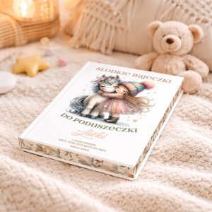 Książka zatytułowana "Słodkie Bajeczki Do Poduszeczki" leży na miękkim kocu, obok pluszowego misia. Na okładce obrazek dziewczynki w kolorowej czapce, przytulającej szarego jednorożca, otoczonej gwiazdkami. W tle widać dekoracyjne elementy, takie jak gwiazdki i małe, pastelowe ozdoby.