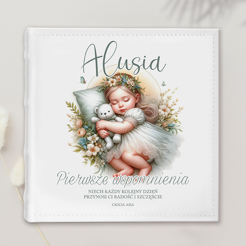 Ilustracja przedstawiająca małą dziewczynkę o blond włosach, uśmiechniętą, śpiącą w białej sukience, przytulającą pluszowego misia. W tle kwiatowa kompozycja z liśćmi i motylami. Na górze napis "Alusia", a poniżej tekst "Pierwsze wspomnienia" oraz życzenia dotyczące radości i szczęścia.