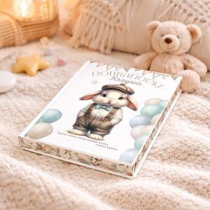Książka z ilustracją małego króliczka w kapeluszu i ubranku na tle pastelowych balonów, leżąca na pluszowym kocu w przytulnym otoczeniu, obok pluszowego misia.