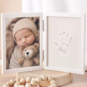 Noworodek śpi w otoczeniu pluszowego misia, a obok znajduje się ramka z odciskiem dłoni i zdjęciem dziecka.