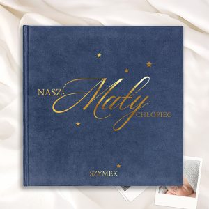 album-welurowy-na-narodziny Nieco otwarta książka w odcieniu granatowym z złotym napisem "NASZ Mały CHŁOPIEC" oraz imieniem "SZYMEK", na tle lnianego materiału i zdjęć polaroidowych na stole.
