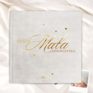 Okładka albumu w kolorze szarym z napisem złotymi literami 'Nasza Mała Dziewczynka' oraz imieniem 'Hania', otoczona małymi, złotymi gwiazdkami, leżąca na jasnym tle z falującymi materiałami.