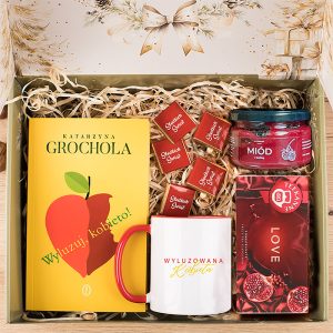 Zestaw podarunkowy zawierający książkę "Wyluzuj, Kobieto!" Katarzyny Grocholi, biały kubek z czerwonym uchwytem i napisem "WYLUZOWANA KOBIETA", słoik miodu, opakowanie herbaty o smaku granatu oraz kilka kwadratowych czekoladek z napisem "Słodkich Świąt" na tle wypełnionym szarym materiałem wypełniającym.