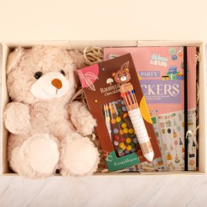 Zestaw w białym pudełku zawierający pluszowego misia w jasnoróżowym kolorze, zestaw kolorowych długopisów, paczkę słodyczy oraz dwa zeszyty z kolorowymi naklejkami i rysunkami.