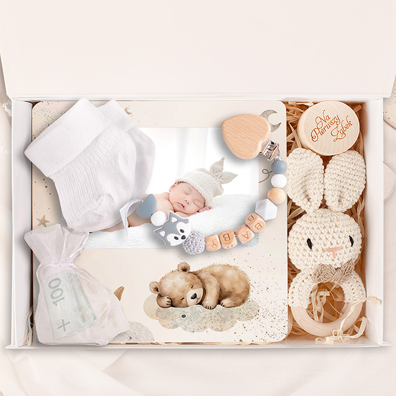 Zestaw prezentowy dla niemowlęcia, zawierający białe skarpetki, zdjęcie śpiącego dziecka w kapeluszu, kolorowy gryzak z motywem zwierząt, koralikową zawieszkę z imieniem, makatkę z ilustracją misia oraz woreczek z banknotem.